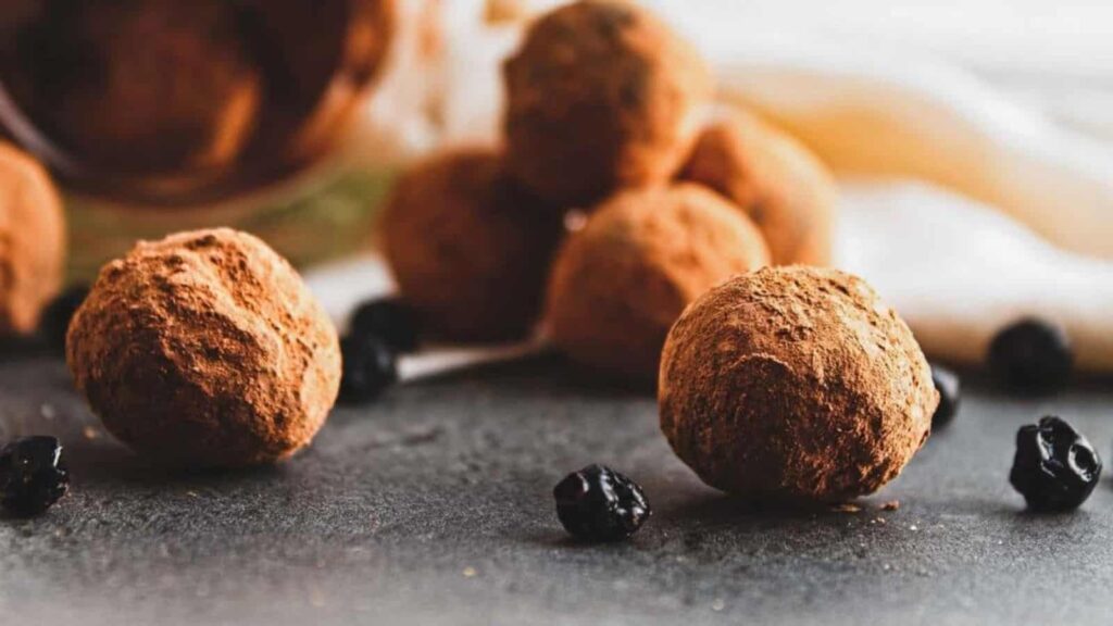 Chocolate truffles coated with cocoa powder are placed on a gray surface. Some of the truffles and small dried berries are in the background, slightly out of focus. A light fabric is draped behind them.