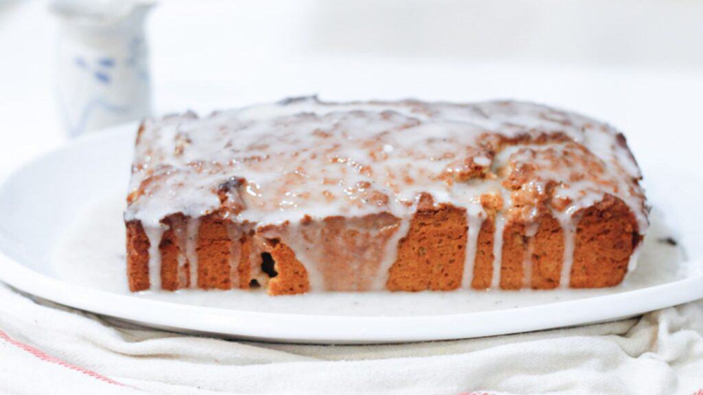 A moist, golden spiced apple butter cake topped with a glossy vanilla glaze drips enticingly down the sides on a white serving plate.