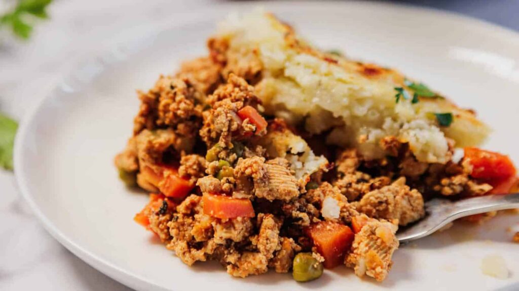 A serving of shepherd's pie with ground meat, peas, carrots, and mashed potatoes on a white plate.