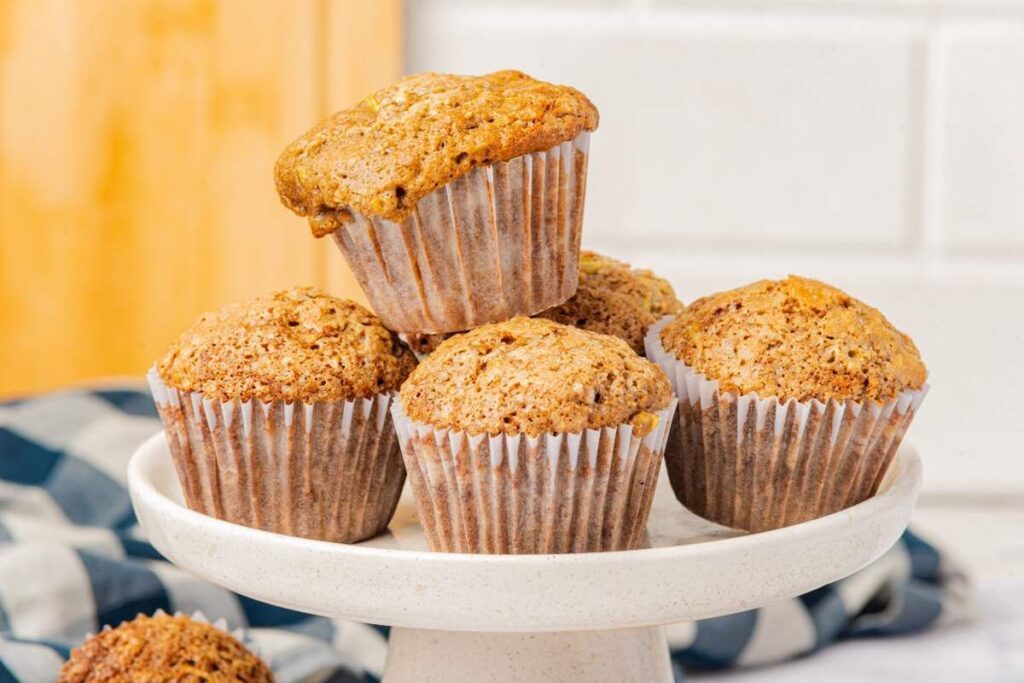 A group of five zucchini muffins is displayed on a white pedestal stand, with a checkered cloth in the background.