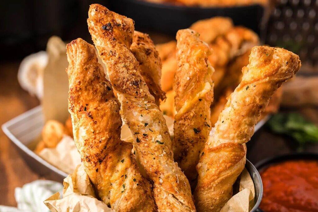 Golden, twisted breadsticks made from puff pastry cheese straws and seasoned with herbs, Parmesan, and mozzarella sit upright in a paper-lined container on a wooden table.