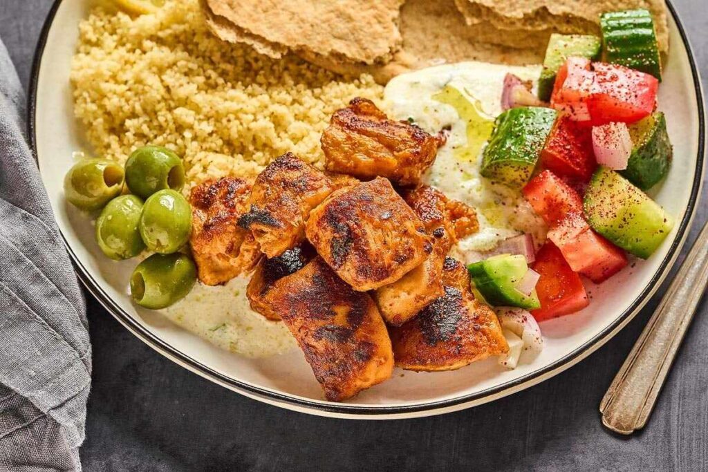 A plate of Aleppo Pepper Chicken Bites, olives, couscous, pita bread, tzatziki, and a cucumber-tomato salad.