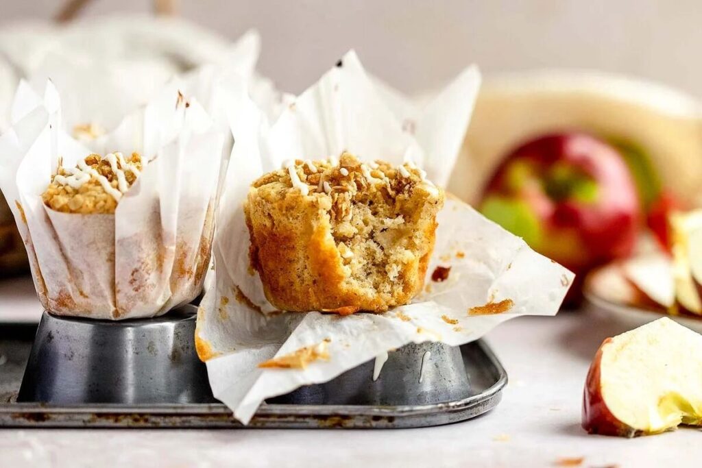 A muffin with a bite taken out sits in a muffin tin, surrounded by apple slices and another muffin.