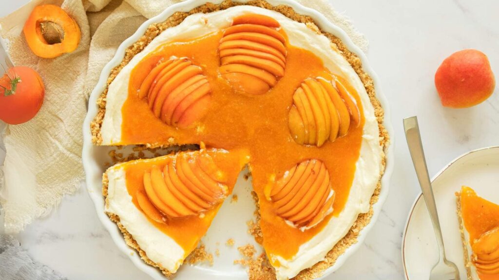 Apricot cheesecake with a creamy base, topped with sliced fresh apricots and apricot glaze, missing one slice on a white plate nearby.