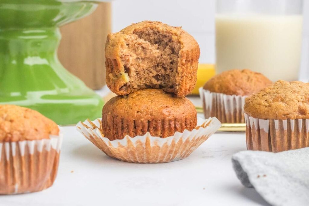 A bitten banana muffin stacked on another muffin, surrounded by more banana muffins, with a glass of milk in the background.