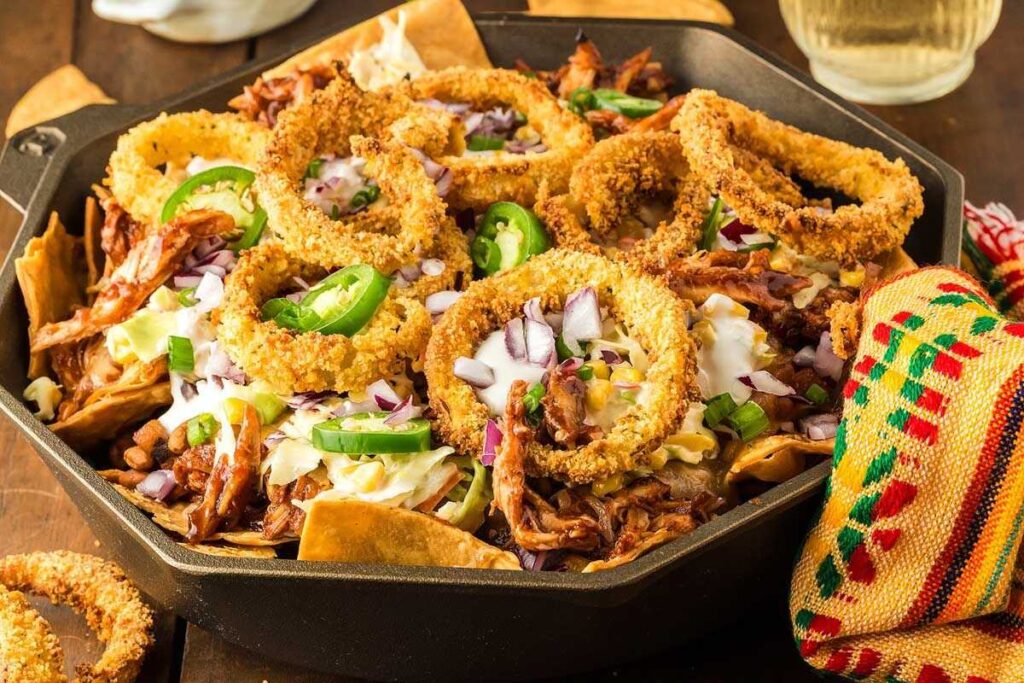  A black cast-iron skillet filled with BBQ chicken nachos topped with onion rings, shredded meat, coleslaw, beans, jalapeno slices, red onions, and melted cheese, placed on a wooden surface with a colorful fabric napkin beside it.