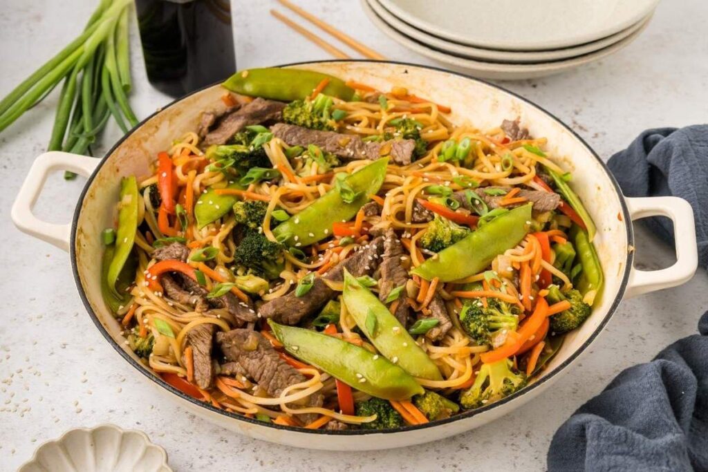 A large pan filled with beef lo mein—stir-fried noodles, tender beef strips, snap peas, broccoli, and carrots—garnished with green onions. 
