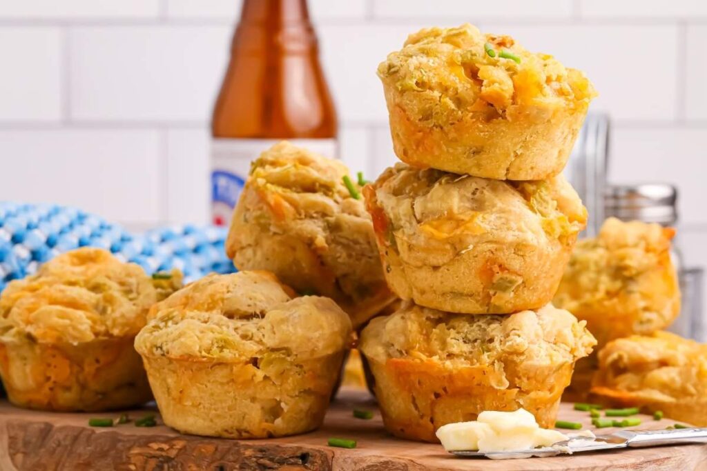 Beer cheese muffins stacked on a wooden board, accompanied by butter and chives.
