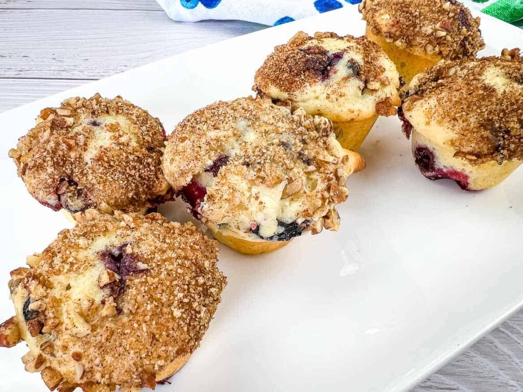 Six freshly baked muffins topped with crumb topping and visible blueberries, arranged on a white rectangular plate against a wooden background.