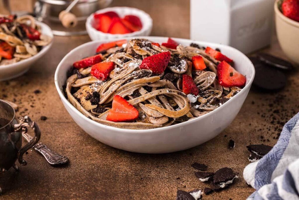 A bowl of Chocolate Pancake Spaghetti topped with strawberries and cookie crumbles sits on a rustic table, inspired by the latest Tik Tok Recipe trend.
