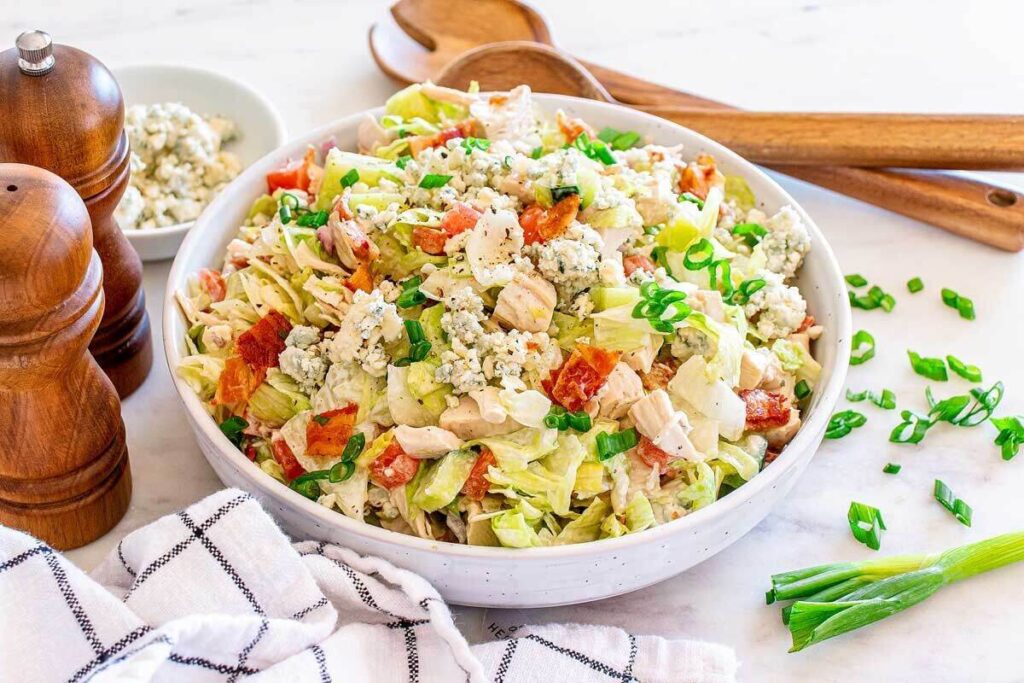  A bowl of chopped chicken blue cheese salad with crisp lettuce, chopped chicken, tomatoes, onion, savory bacon, tangy blue cheese crumbles, and fresh green onions. Wooden salad utensils, a pepper mill, and a cloth napkin are nearby.