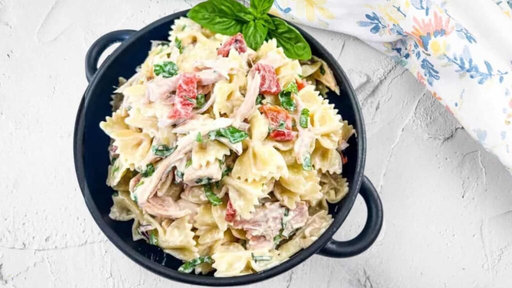 Overhead shot of a black bowl filled with creamy pasta salad, featuring farfalle pasta, chicken, red bell peppers, and fresh basil, garnished with a basil sprig, against a white textured background with a floral patterned cloth in the corner.