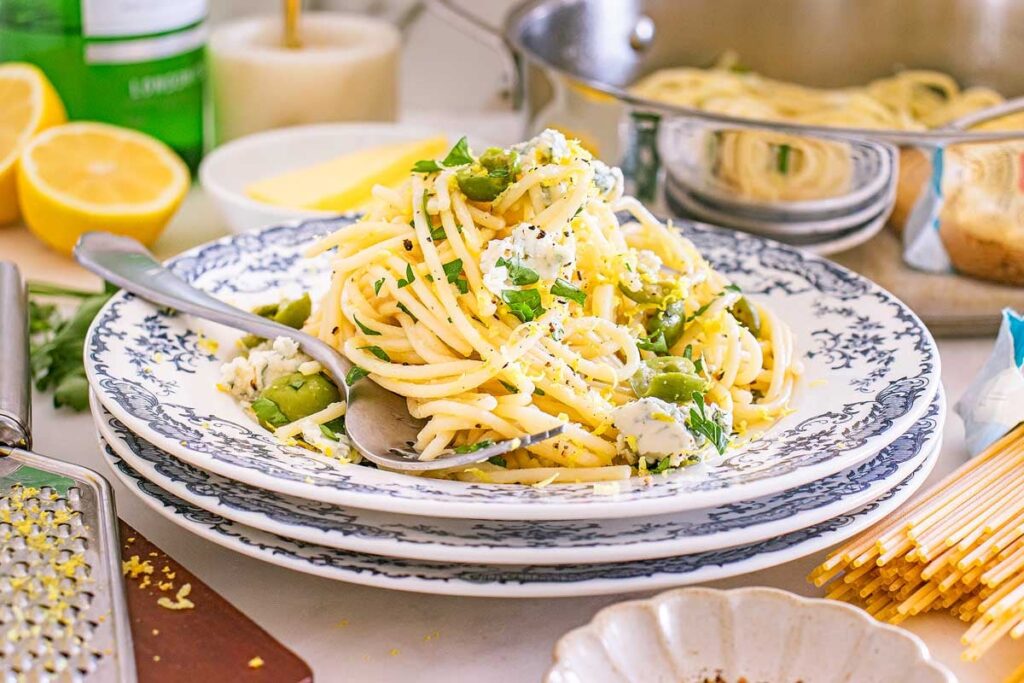 A plate of dirty martini pasta with green vegetables and cheese, served with a spoon and surrounded by fresh cooking ingredients.
