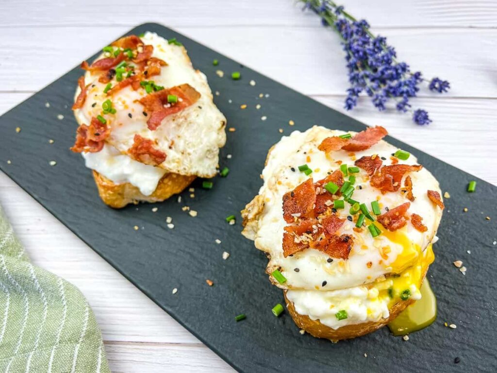 Two open-faced egg sandwiches on a black slate, each with a fried egg on top of a layer of white spread and toasted bread, garnished with bacon bits, chives, and everything bagel seasoning, with a sprig of lavender in the background.