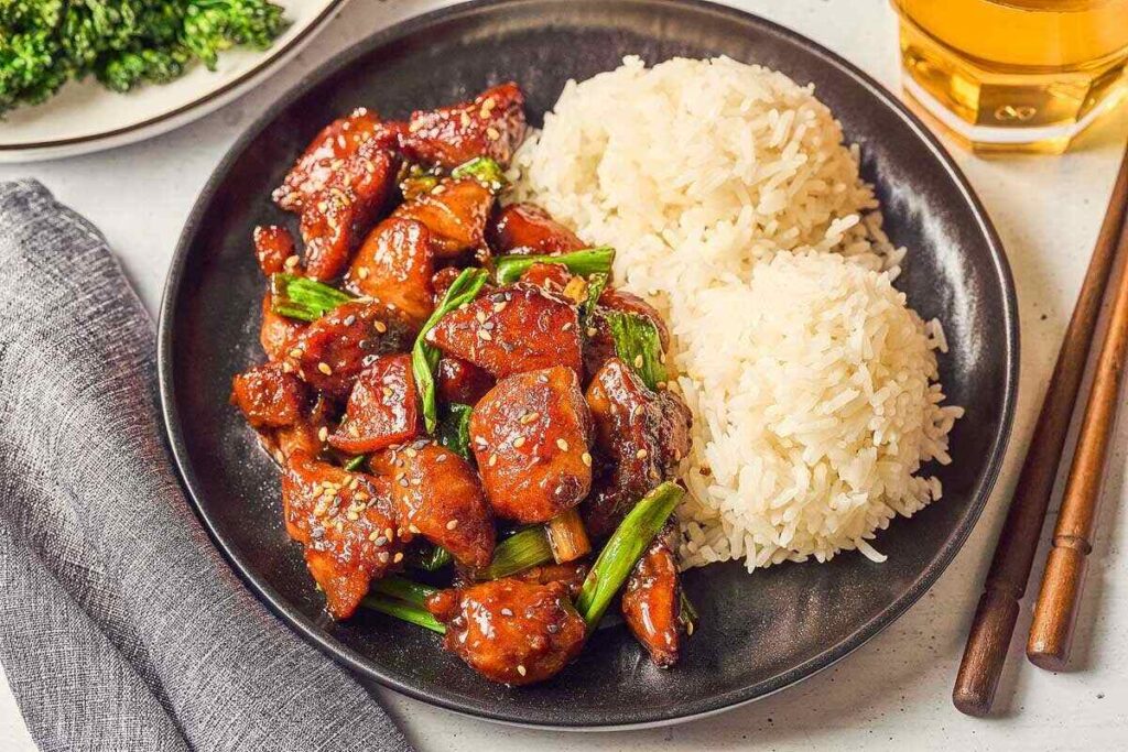 A plate featuring Five Spice Chicken Stir Fry with glazed chicken, green onions, and sesame seeds sits invitingly beside two servings of white rice. 