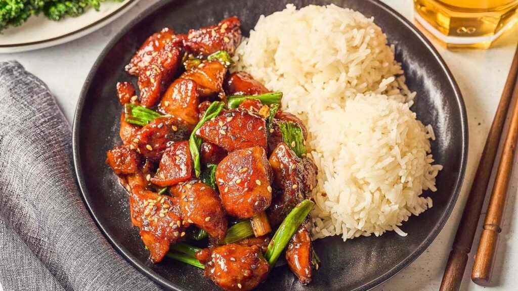 Diced cooked chicken pieces mixed with sliced green onions and sesame seeds are shown on a plate alongside two servings of white rice.