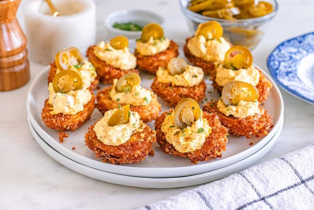 Plate of fried deviled eggs topped with creamy spread and sliced jalapeños, placed on a white plate, with a striped cloth and bowls in the background.