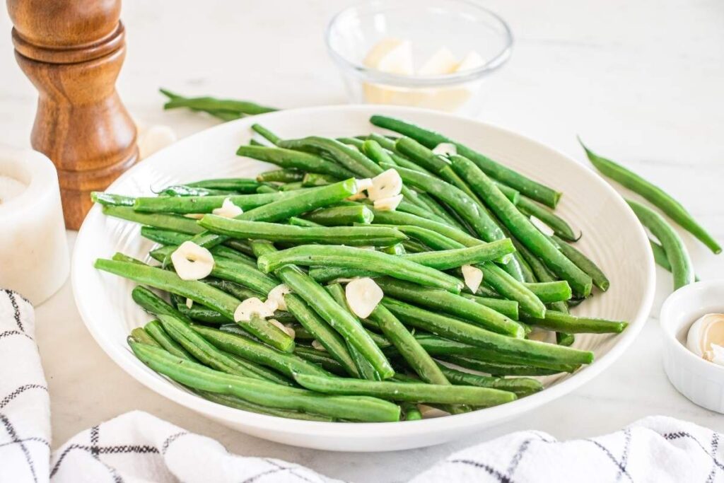 A white plate of garlic green beans is topped with sliced garlic, next to a wooden pepper mill and a bowl with chunks of butter.
