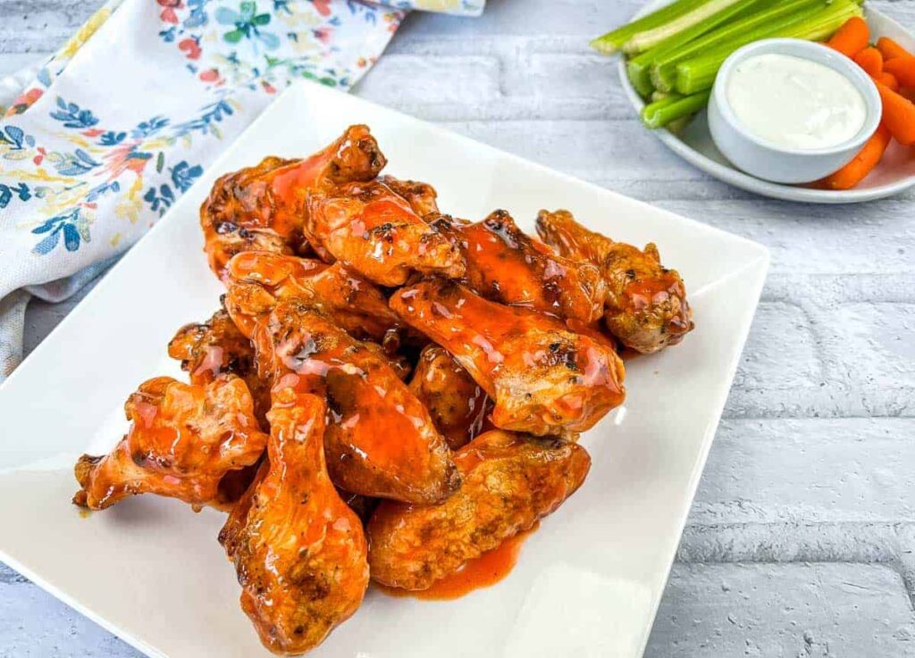 A square white plate filled with saucy buffalo chicken wings sits on a white surface, with celery, carrots, and ranch dip in the background.