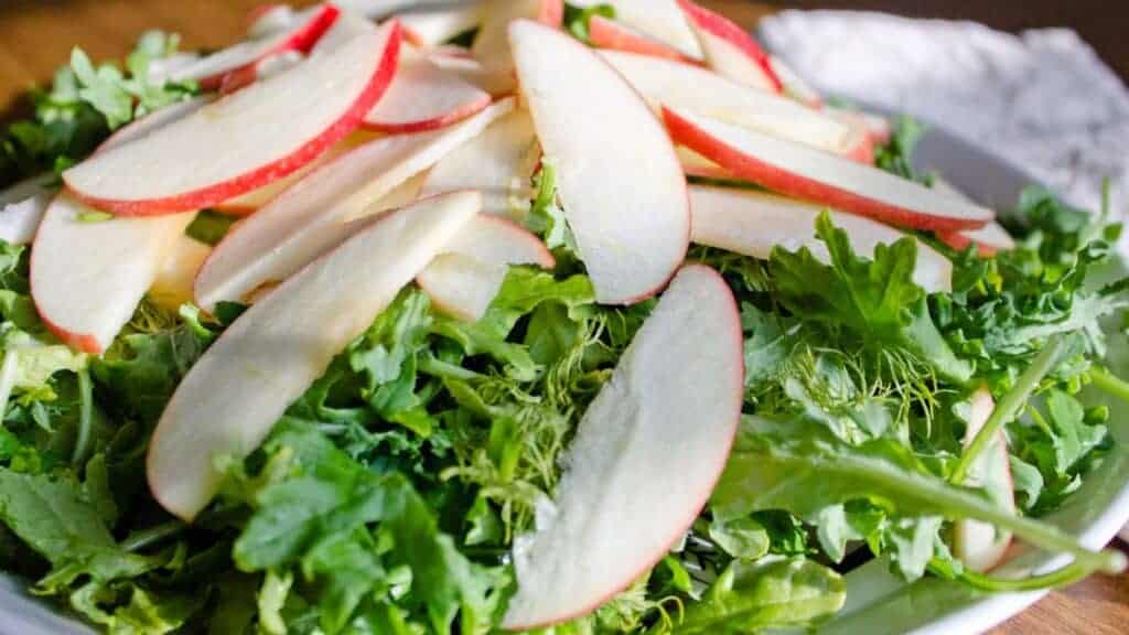 A delightful apple and greens salad featuring fresh kale adorned with thin slices of crisp red apple rests on a wooden surface, complemented by a glimpse of white cloth in the background.