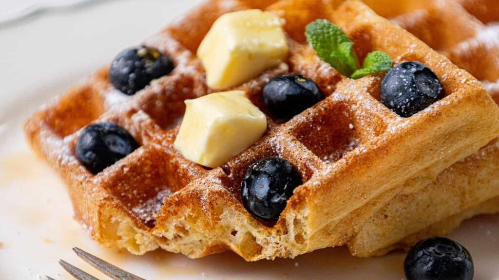 Close-up of golden brown waffles topped with blueberries, butter cubes, a sprinkle of powdered sugar, and a small mint leaf. A fork is partially visible in the foreground.