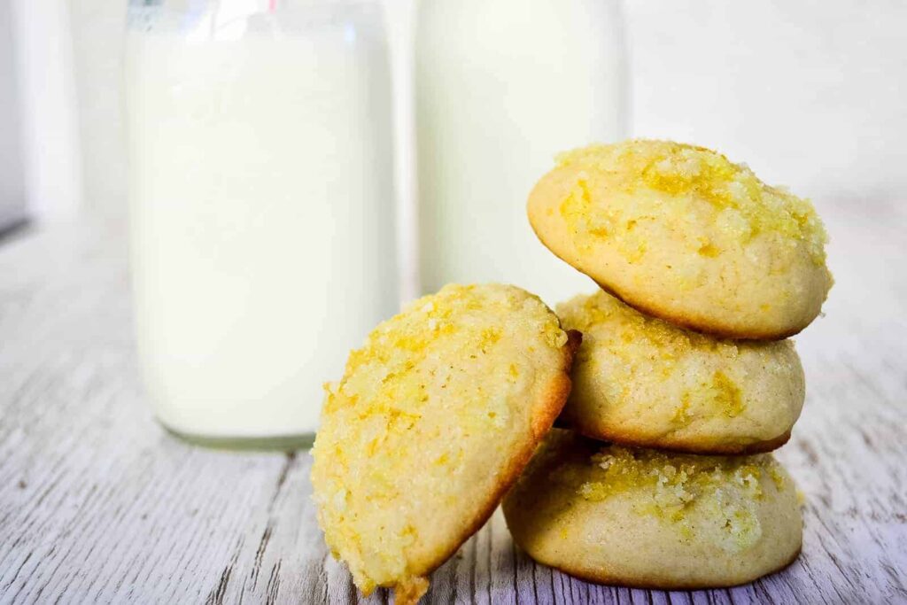 A single cookie leaning on a stack of Lemon Sugar Cookies in front of two bottles of milk.