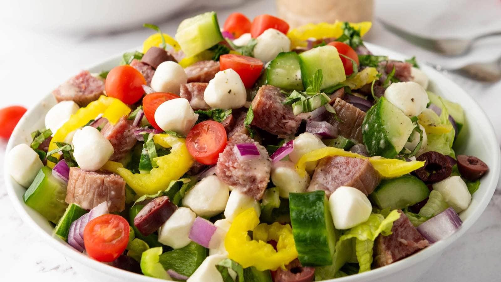 Italian salad with lettuce, cherry tomatoes, cucumber, red onion, mozzarella balls, olives, pepperoncini, and sliced meat.