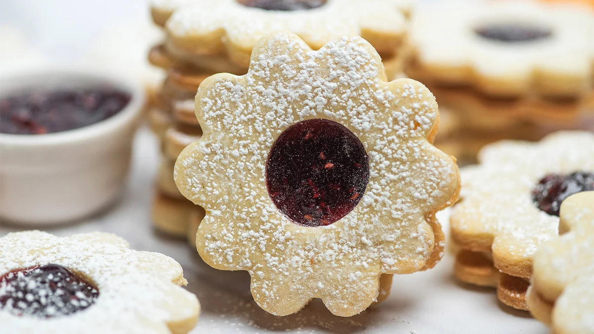Stacked sandwich cookies filled with buttercream and red jam, dusted with powdered sugar on top.