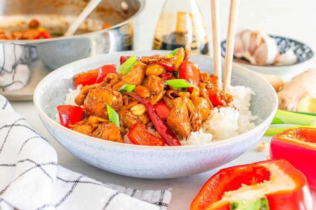A bowl of kung pao chicken features stir-fried chicken with red peppers, peanuts, and green onions on rice, accompanied by chopsticks.