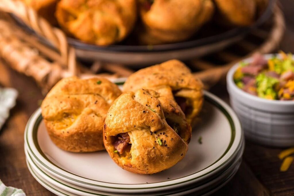 Three savory ham and broccoli crescent cups, with diced toppings in a small bowl nearby.