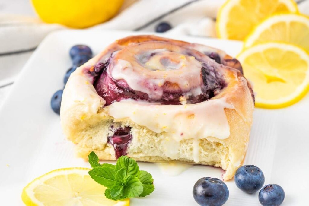  A lemon blueberry sweet roll with icing, garnished with fresh blueberries and lemon slices, sits invitingly on a white plate.