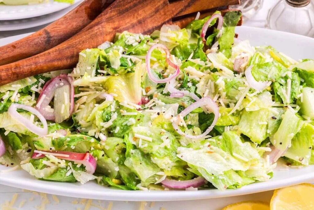 A close-up of a fresh Lemon parmesan chopped salad with shredded romaine, onions, grated cheese, and wooden serving utensils on a white dish.