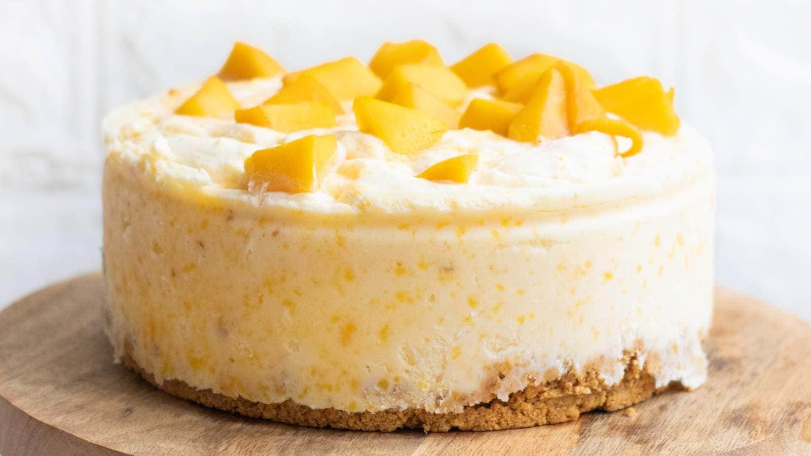 A slice of creamy cheesecake with a crumbly crust, topped with chunks of fresh mango, sits on a white plate with a fork.