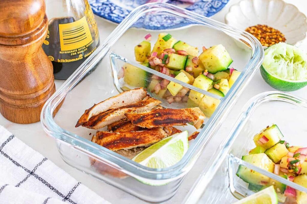 Glass meal prep container with Thai chicken slices, cucumber salsa, and a lime wedge on a table.