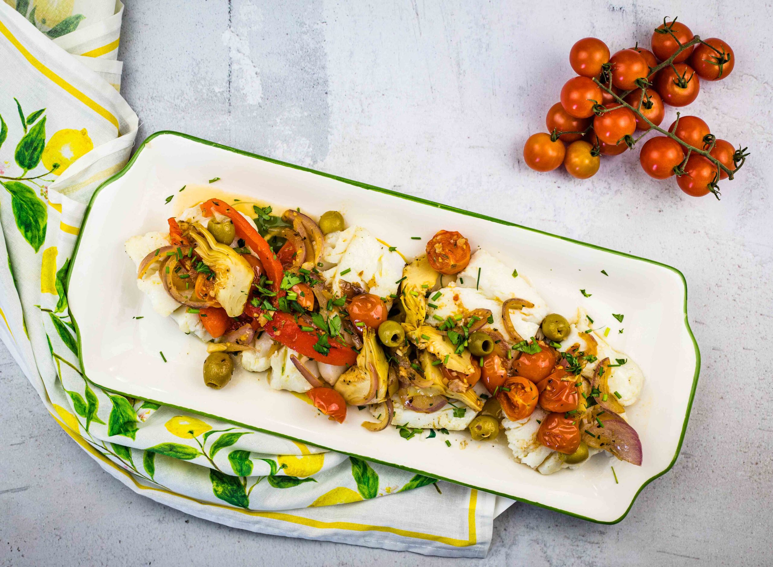 A rectangular plate with Mediterranean Cod placed on a lemon-patterned cloth. A vine of cherry tomatoes is beside the plate.