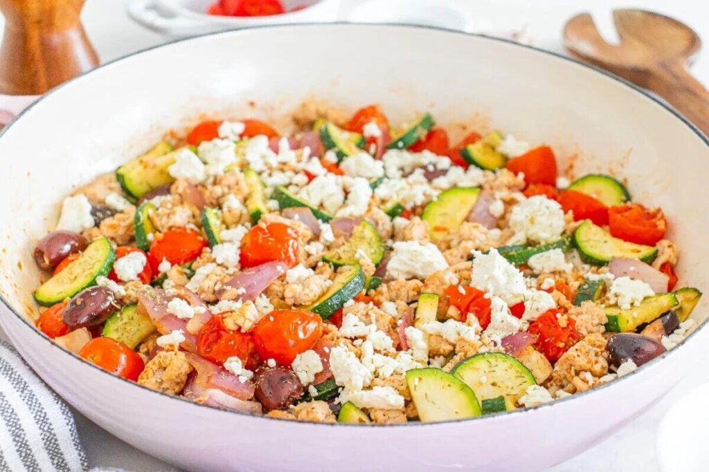 A skillet filled with a mixture of ground turkey, zucchini, cherry tomatoes, red onions, olives, and crumbled feta cheese.
