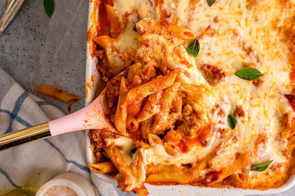 Baked mostaccioli pasta with melted cheese and fresh basil, served with a spoon from a casserole dish.