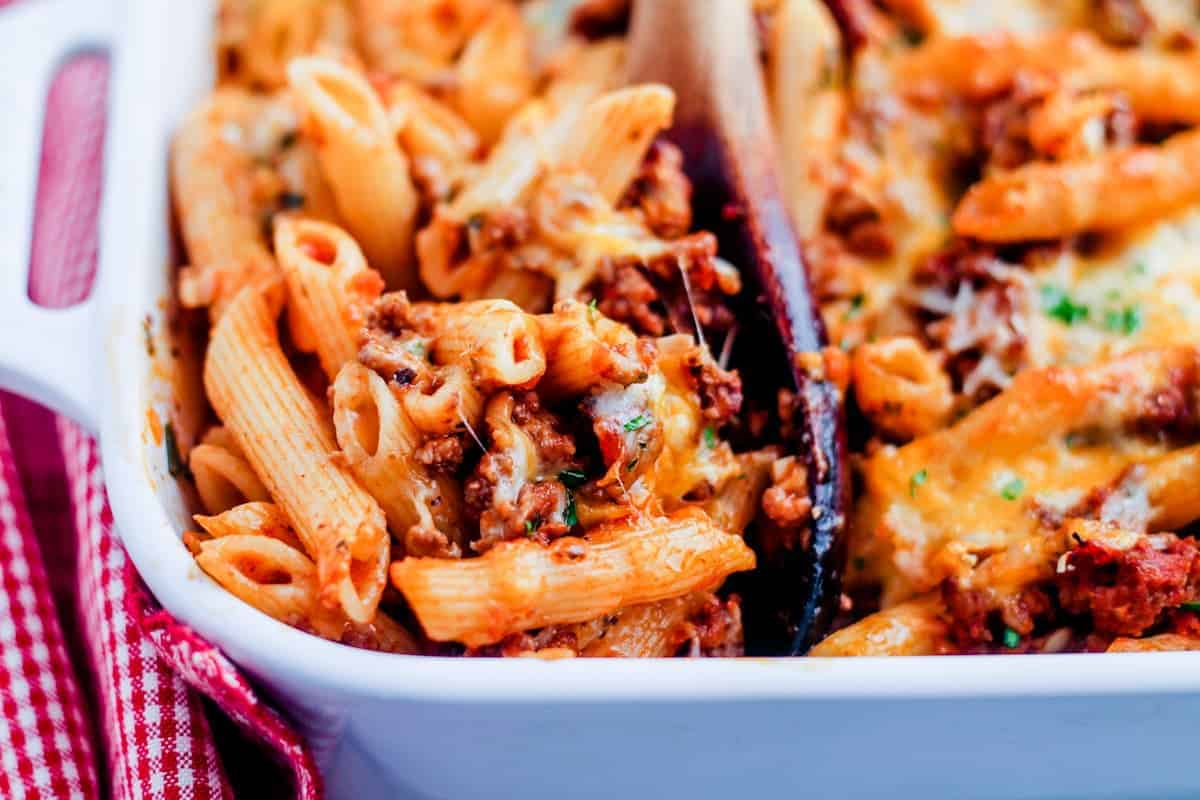 A wooden spoon scooping a portion of penne pasta bake.