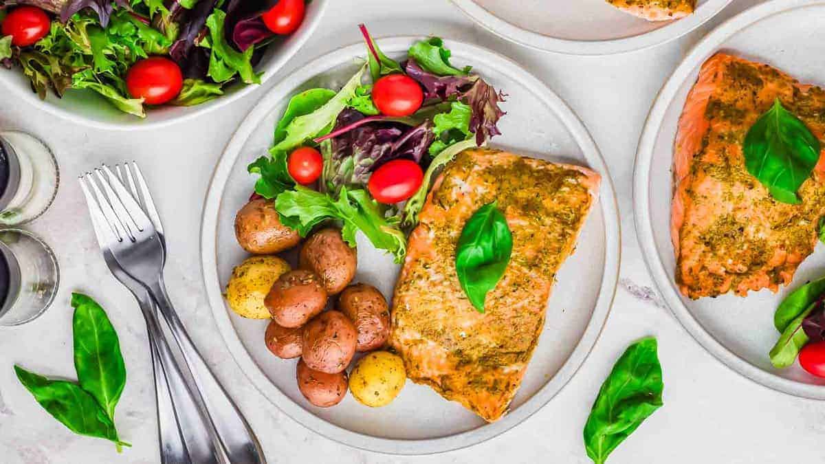 Baked salmon fillet topped with green pesto sauce, served on a white plate with roasted vegetables and herbs.