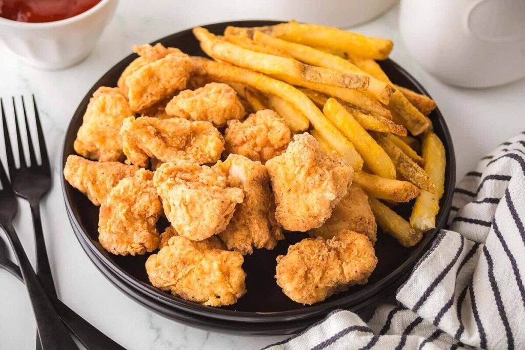 A black plate with crispy popcorn chicken and golden French fries, next to a striped napkin and utensils.