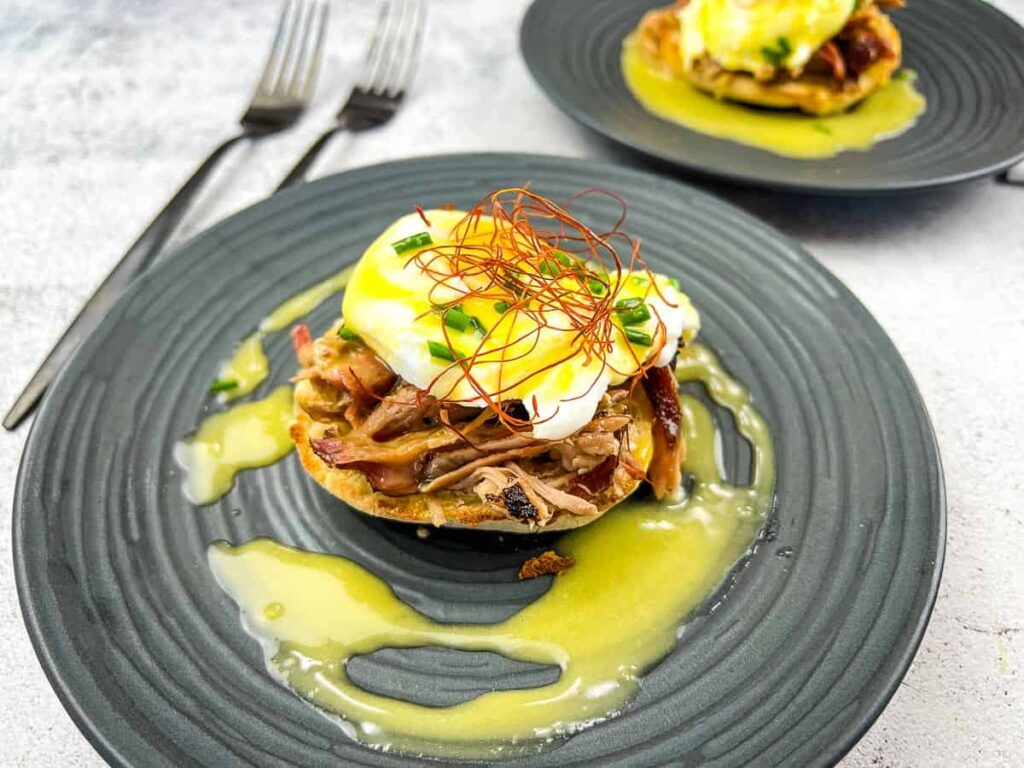 Eggs Benedict dish on a gray plate featuring a toasted muffin topped with pulled meat, a poached egg, chili threads, and chives, drizzled with hollandaise sauce. A second plate with a similar dish is in the background, with two forks on the left side.