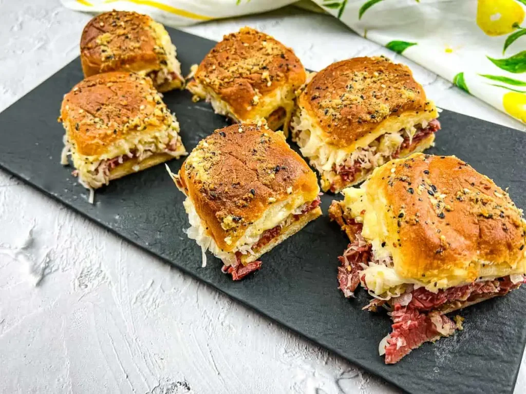 Six Reuben sliders with toasted, seeded buns, stacked with corned beef, Swiss cheese, and sauerkraut, arranged on a dark slate board on a white textured surface.