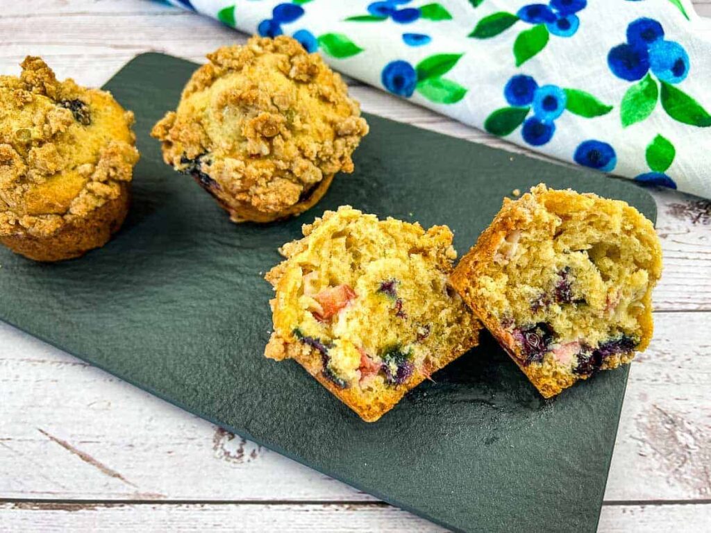 Three blueberry muffins on a dark green slate surface with a cut muffin revealing blueberries and crumble topping, a white napkin with blue and green blueberry designs rests nearby on a light wooden table.