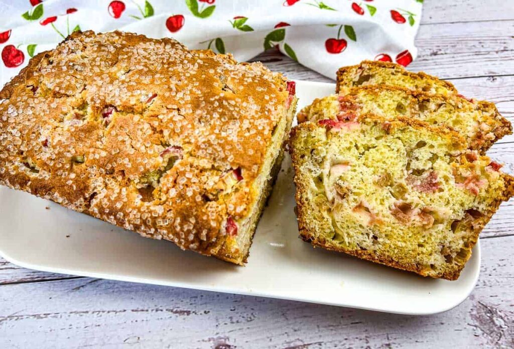 A partially sliced loaf of rhubarb bread on a white rectangular plate, alongside three slices. The bread has a golden-brown, sugared crust and visible pieces of rhubarb. A cherry-patterned cloth is in the background.