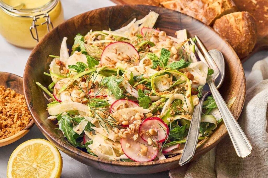 A fresh shaved asparagus salad with radishes, fennel, herbs, and nuts in a wooden bowl, served alongside crusty bread and a lemon wedge.