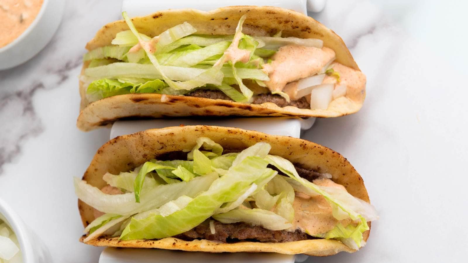 Two tacos filled with ground beef, shredded lettuce, diced onions, and a creamy sauce.