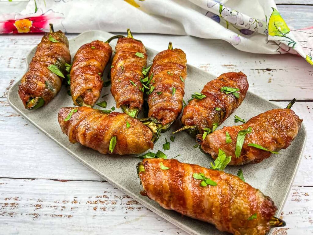 A rectangular plate with eight bacon-wrapped jalapeño poppers, garnished with chopped parsley, on a white wooden table with a patterned cloth in the background.