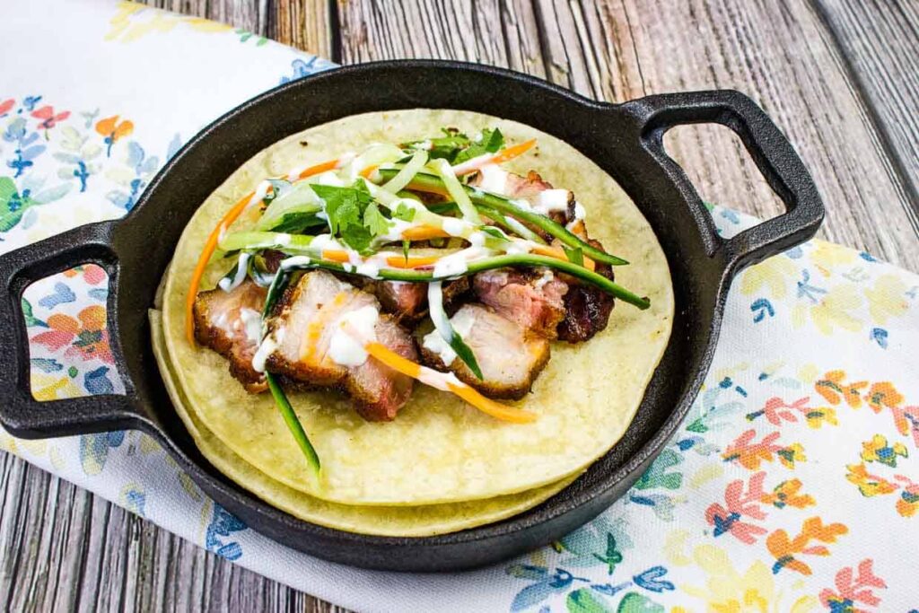 Close-up of a loaded pork belly taco served in a black cast iron skillet on a floral napkin, featuring a stack of two tortillas topped with sliced, crispy pork belly, shredded carrots, green onion slivers, cilantro, and drizzled with a white sauce.