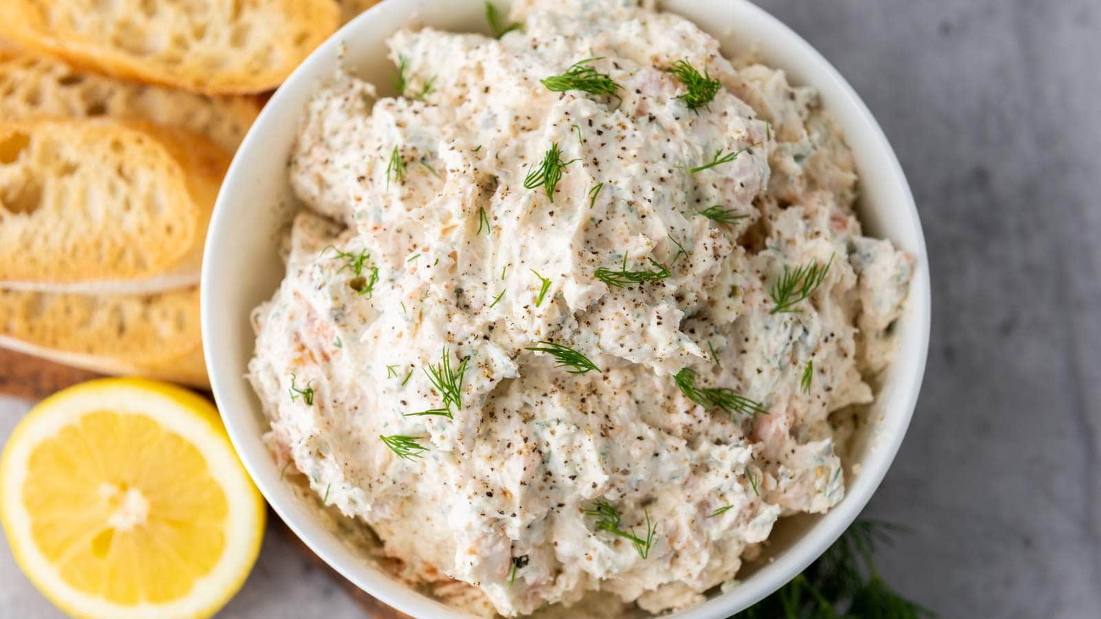 A bowl of smoked salmon spread garnished with fresh dill and black pepper, with sliced baguette and a lemon wedge on the side.