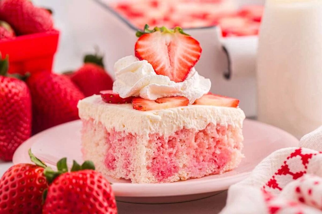 A slice of strawberry poke cake, topped with whipped cream and garnished with sliced strawberries, sits elegantly on a plate surrounded by fresh strawberries.